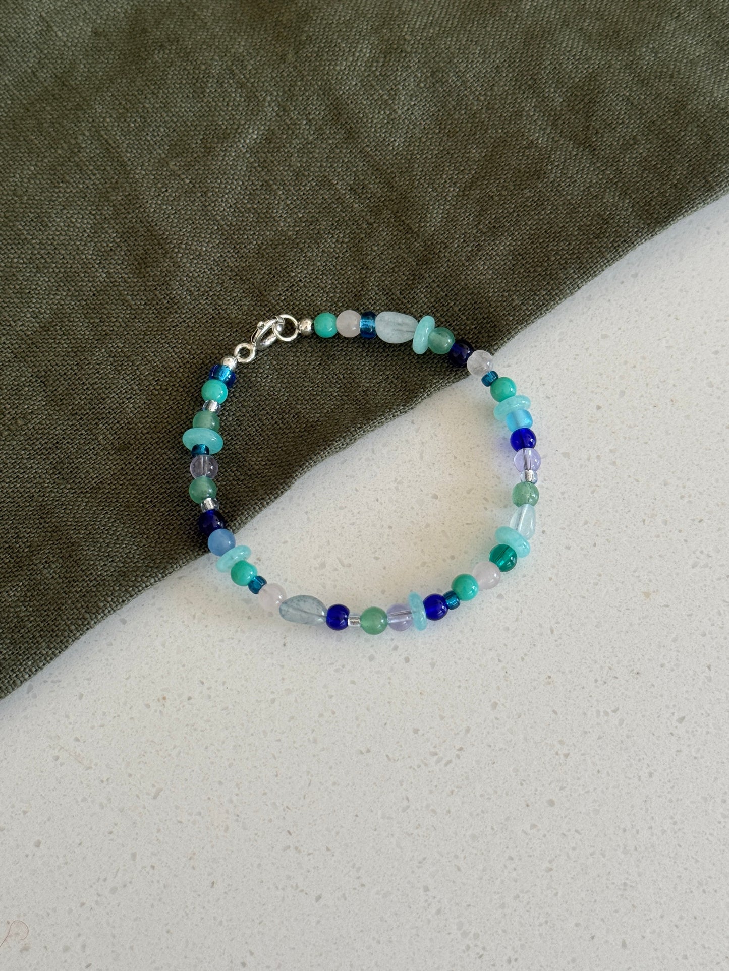A beaded bracelet with small blue, green, and light aqua beads, featuring a silver clasp, displayed on a split background of dark olive green fabric and a light gray textured surface.