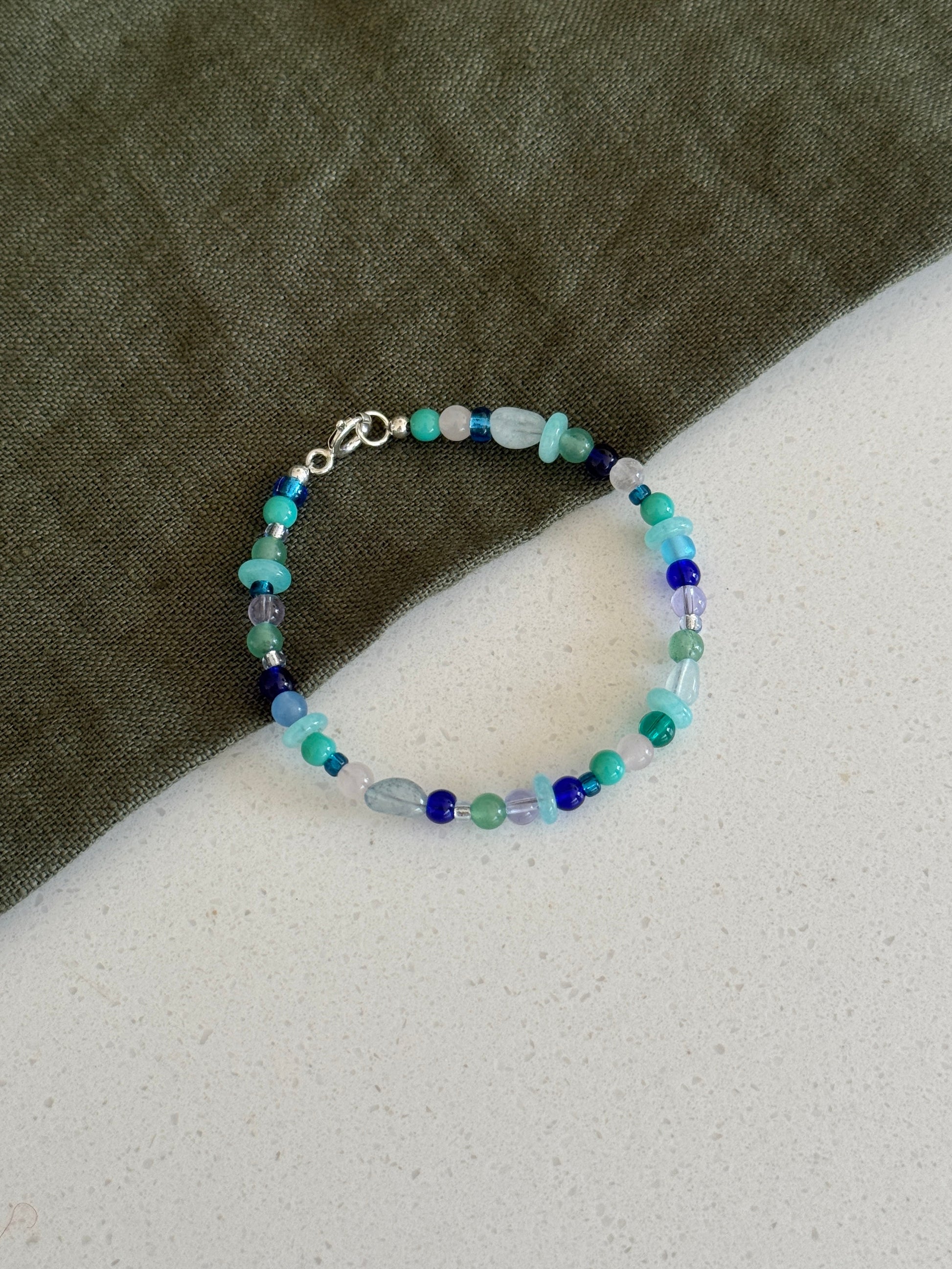A beaded bracelet with small blue, green, and light aqua beads, featuring a silver clasp, displayed on a split background of dark olive green fabric and a light gray textured surface.