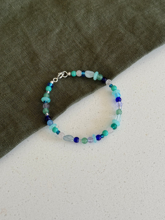 A beaded bracelet with small blue, green, and light aqua beads, featuring a silver clasp, displayed on a split background of dark olive green fabric and a light gray textured surface.